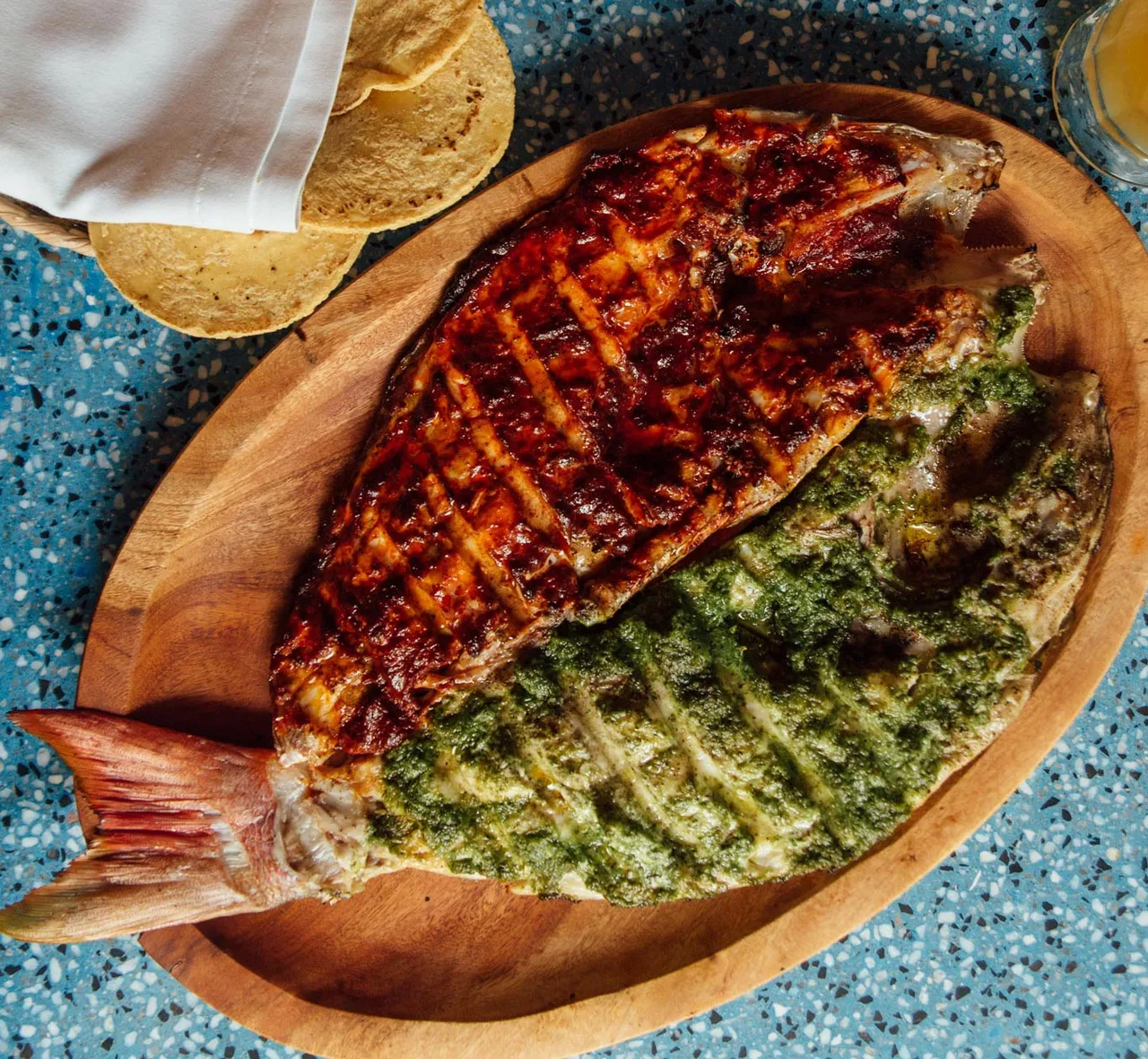 A whole fish grilled in two halves, one red and one green, on a wooden board.